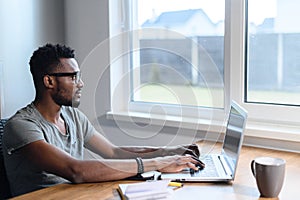 Smart African-American guy wearing glasses and casual t-shirt using laptop