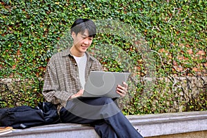 Smart Asian male college student remote working in the city park, using his laptop on a bench