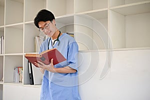 Smart Asian male medical student wearing glasses and reading a book while standing in library