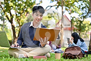 Smart Asian male student reading a book, using laptop, doing homework in the campus`s park