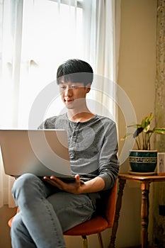 Smart Asian man focusing on his work on laptop, using laptop on a chair in his living room