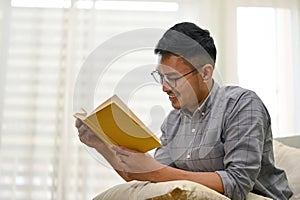 Smart Asian man wearing glasses reading a book on sofa in his living room. side view