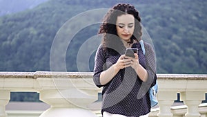 Smart, beautiful brunette corrects curly hair and prints sms on the phone at against the backdrop of outstanding