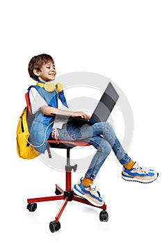 Smart blonde boy sitting on red chair and looking on notebook display over white background. Back to School.