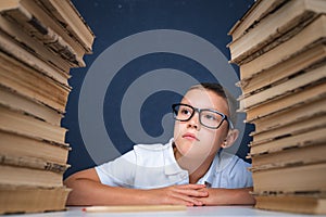 Smart boy in glasses sitting between two piles of books and look away thoughtfully