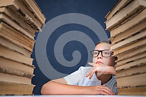 Smart boy in glasses sitting between two piles of books and look away thoughtfully