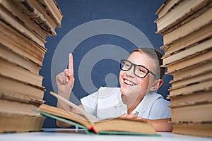 Smart boy in glasses sitting between two piles of books and look up, pointing finger