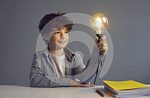Smart boy pupil sitting at desk holding glowing light bulb studio portrait