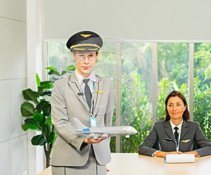 Smart caucasian woman smiles, stands, holds airplane model, wear pilot hat, looking to camera with business women and space at