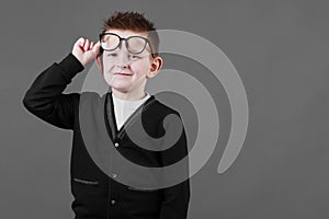 Smart child boy adjusts his glasses and having fun on the grey background, close-up. Studio juvenile portrait in casual