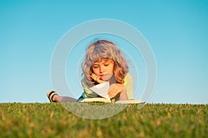 Smart child reading book, laying on grass in field on sky and grass field. Portrait of clever kids.