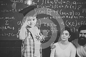 Smart child, wunderkind in graduate cap pointing at his head. Boy presenting his knowledge to mom and dad. Parents