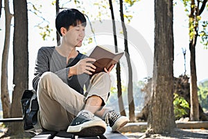 Smart and chilling young Asian male college student reading a book while relaxing in the park