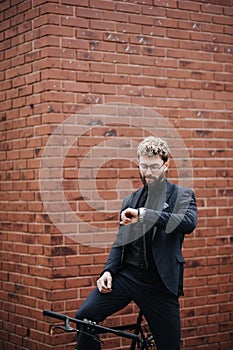 Smart and confident businessman looking at his watch while going to work by bicycle against brick wall background