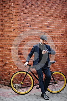 Smart and confident businessman looking at his watch while going to work by bicycle against brick wall background