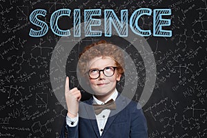 Smart confident kid boy with red hair pointing up at science inscription on chalkkboard