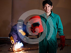 Smart confident young Asian man wearing a factory uniform holding a welding helmet posing smiling to a camera with soft background