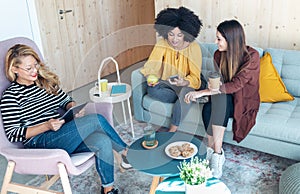 Smart entrepreneur multiethnic women talking while taking a break and showing photos with smart phone sitting on couch at