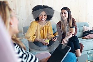 Smart entrepreneur multiethnic women talking while taking a break and showing photos with smart phone sitting on couch at