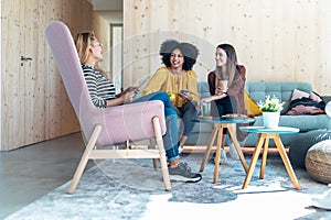 Smart entrepreneur multiethnic women talking while taking a break and showing photos with smart phone sitting on couch at
