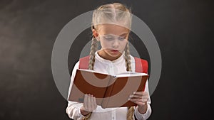 Smart female kid reading book, blackboard on background, thirst for knowledge