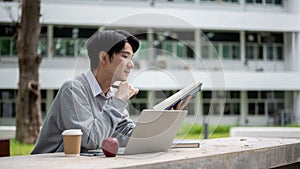 A smart and focused Asian male university student is reading a book in a campus park