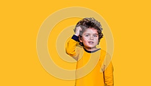 Smart funny curly kid scratches his head while thinking. Little wunderkind ready to go to school isolated on yellow