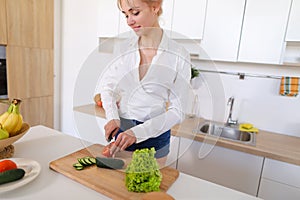 Smart girl slices cucumber with knife and smiles, standing at ki