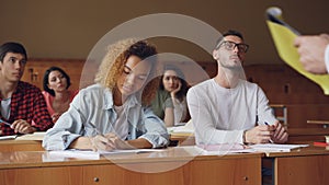 Smart guy in glasses student is talking to teacher sitting at desk in lecture hall with young people around him. Higher