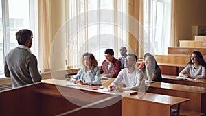 Smart guy student is raising hand and talking to professor while fellow students are listening to them and smiling