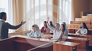 Smart guy student is raising hand and talking to professor while fellow students are listening to them and smiling