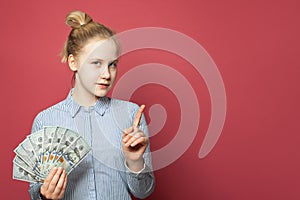 Smart happy cunning young girl showing money us dollars and pointing finger up on pink background