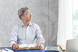 Smart healthy business old man sitting in office, Asian elder thinks looking out of the windows while reading a book