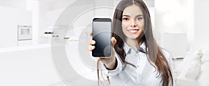 smart home control concept smiling woman showing cell phone screen on kitchen and living blurred background