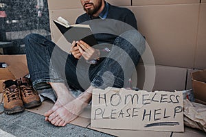 Smart homeless man is sitting on cardboard and reading a book. He is very concentrated on that. There is a sigh which