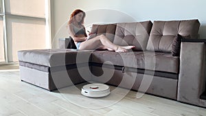 Smart House. Cleaning the house`s robotic vacuum cleaner, when women based on sofas play mobile phones