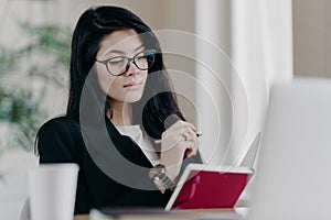 Smart intelligent female teacher in glasses for vision correction learns online courses, makes notes in notepad as watches webinar