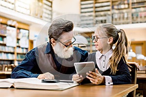 Smart kid girl in eyeglasses sitting at the table in old library and holding tablet, showing something on tablet to her