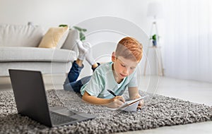 Smart kid laying on carpet with laptop, taking notes