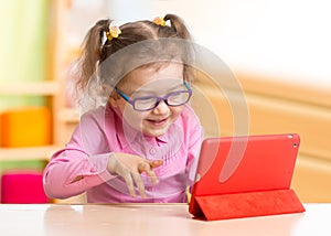 Smart kid in spectacles using tablet PC or e-book sitting at table in her room