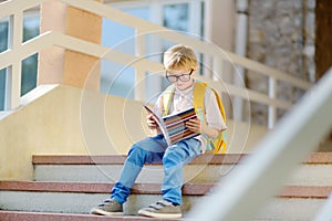 Smart little child sitting and reading on the stairs of school building. Quality education for children. Portrait of funny nerd