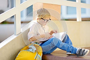 Smart little child sitting and reading on the stairs of school building. Quality education for children. Portrait of funny nerd