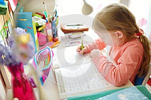 Smart little schoolgirl doing her homework at her table at home. Child learning to write and read