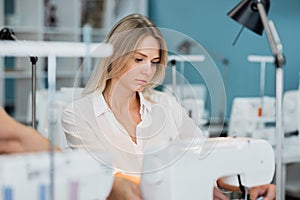 A smart-looking pretty blond woman wearing white shirt is sewing with the electric sewing-machine. Fashion, tailor`s