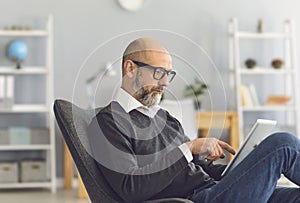 A smart mature man with glasses looks in a tablet in his hands while sitting in a comfortable chair in the living room.