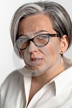 Smart mature woman in white shirt looking thoughtfully at the camera. Pretty Caucasian gray haired model in glasses