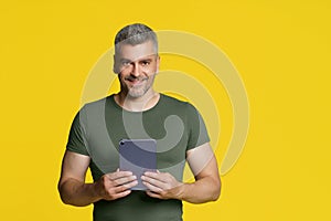 Smart middle aged handsome man holding digital tablet in hands smiling looking at camera isolated on yellow background