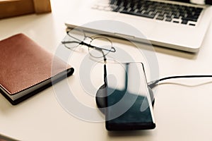 Smart phone being charged on wireless charger alongside with laptop, glasses, and notebook on table.