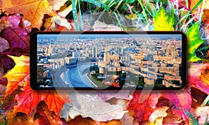 Smart phone with Moscow aerial panorama on colourful autumn leaves fallen onto green grass