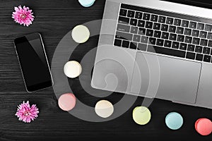 Smart phone, notebook computer and macarons with flowers on black wooden table, top view